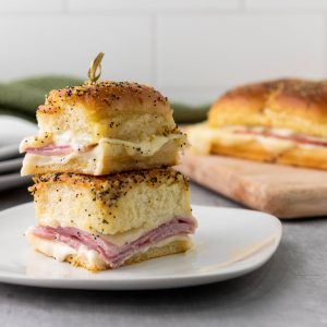 A close-up image of two Ham and Cheese Sliders stacked on a white plate. The sliders are made with soft rolls, ham, cheese, and a creamy sauce.