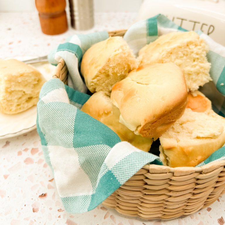 A close-up shot shows a round, woven basket filled with several warm, golden-brown bread rolls, showing their soft texture. The basket is lined with a teal and white checkered cloth napkin. To the left, a single roll sits on a small, white plate with a scalloped edge.