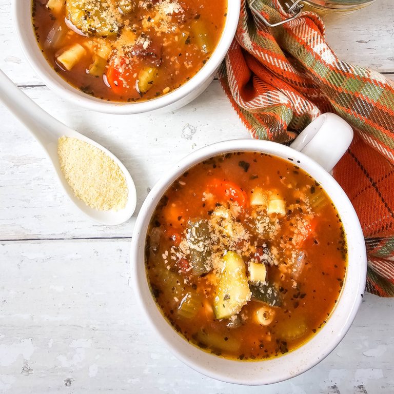 Two bowls of minestrone soup with pasta, vegetables, and parmesan cheese, a spoon with parmesan on the side, and a patterned cloth on a white wooden surface.