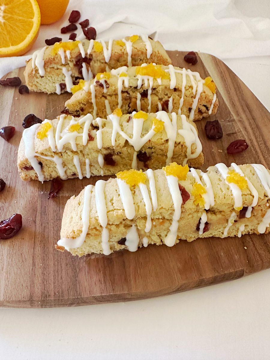 A close-up, slightly angled photo of four finished Orange Cranberry Biscotti on a wooden cutting board. The cookies are topped with white chocolate drizzle and orange zest, with a sliced orange and loose dried cranberries visible in the background.