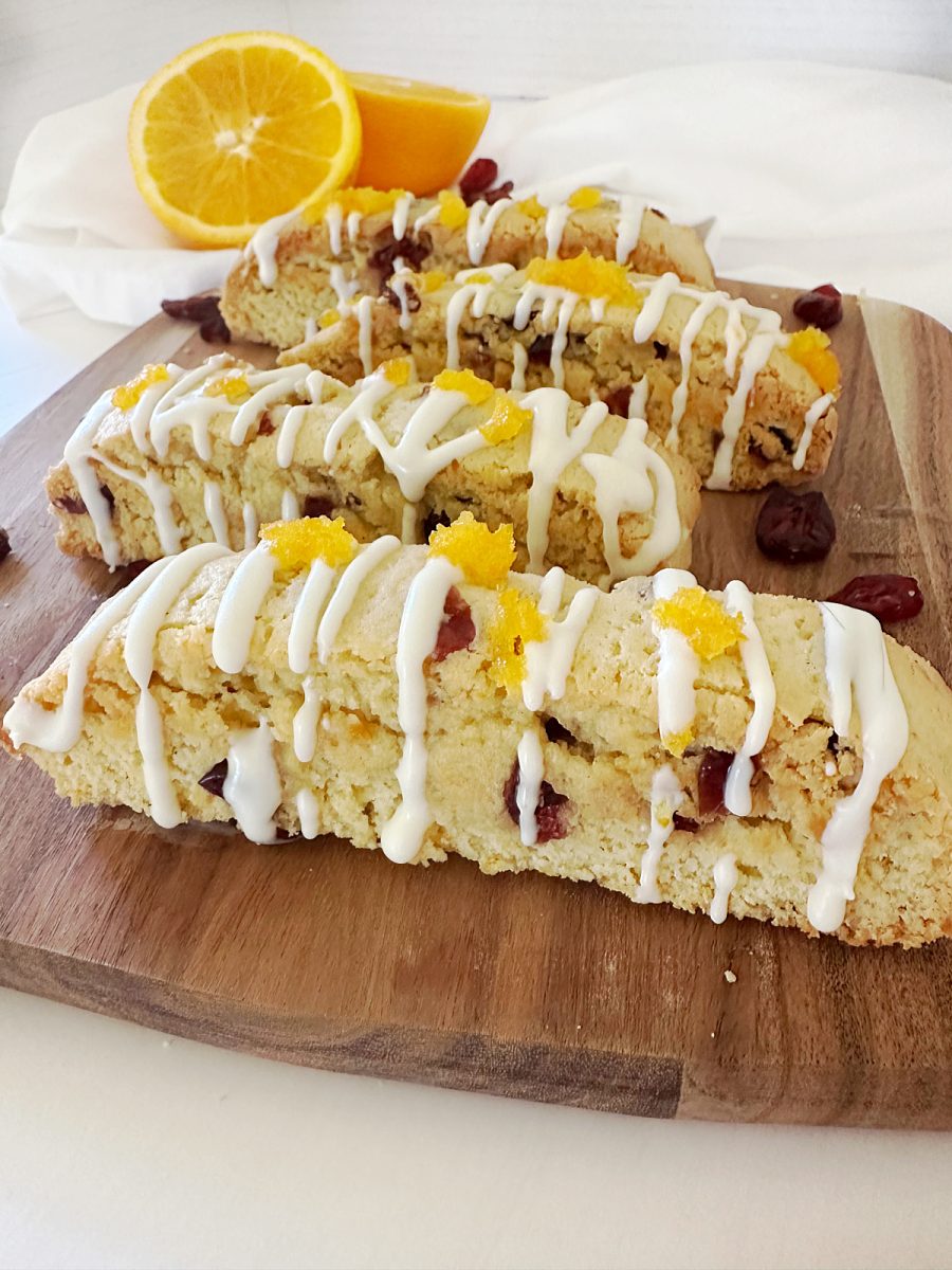 Close-up, angled photo focusing on four finished Orange Cranberry Biscotti on a wooden board. The cookies show the texture, cranberries, white chocolate drizzle, and the bright orange zest garnish. A sliced orange is blurred in the background.