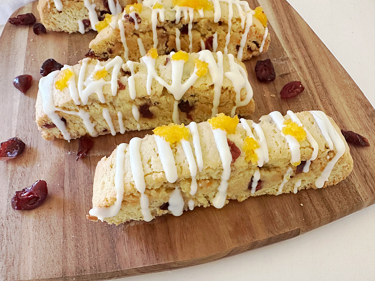 Close-up view of three finished Orange Cranberry Biscotti angled on a wooden cutting board, drizzled with white chocolate and topped with bright orange zest sugar. Scattered dried cranberries surround the cookies.