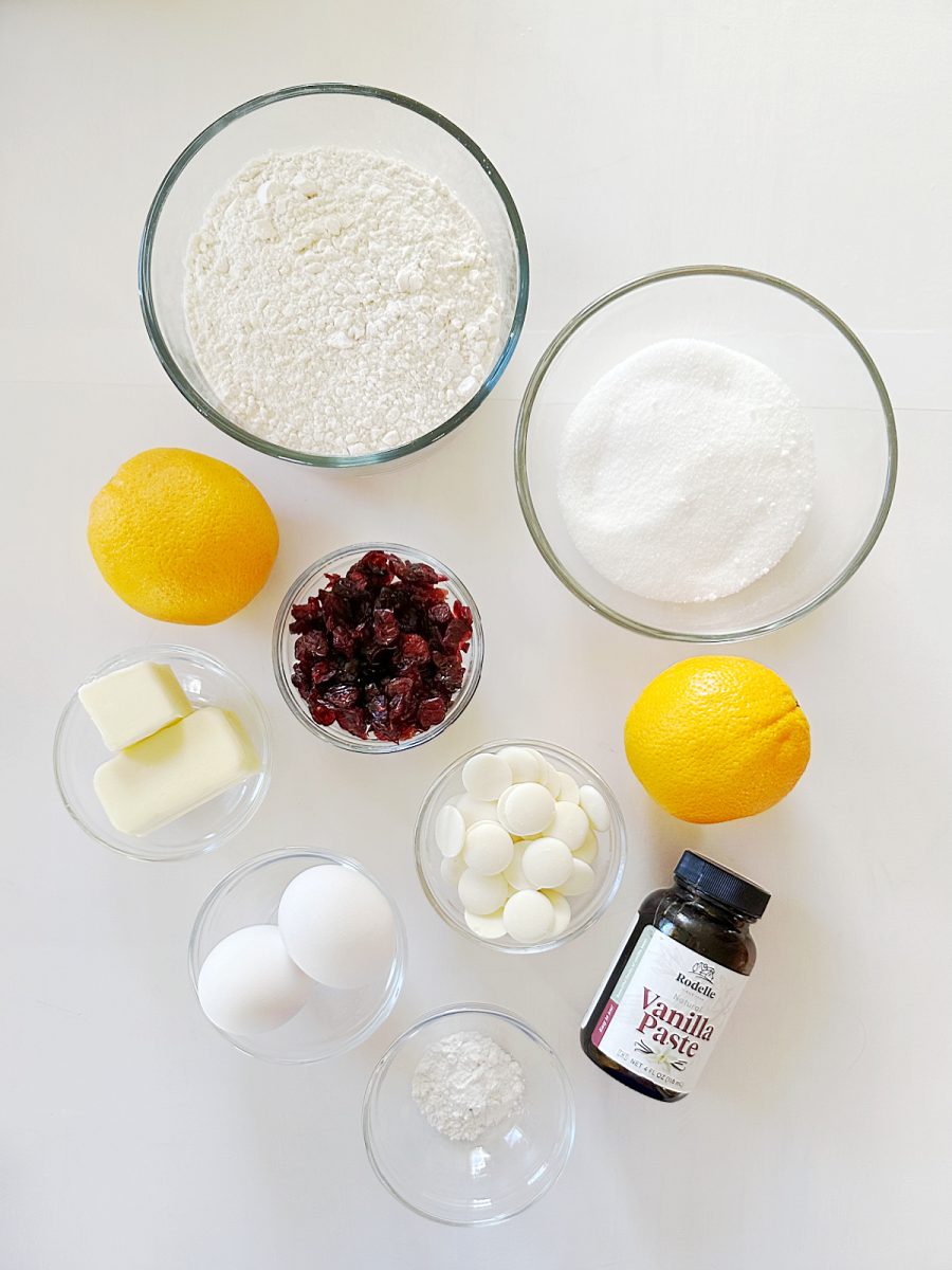 An overhead flat lay of all the Orange Cranberry Biscotti ingredients arranged neatly in clear glass bowls on a white surface. Ingredients include flour, sugar, butter, two eggs, two whole oranges, dried cranberries, white chocolate wafers, vanilla paste, and baking powder.