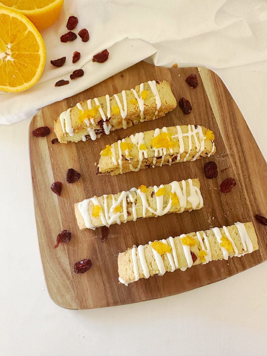 Overhead flat lay photo showing four finished Orange Cranberry Biscotti, each drizzled with white chocolate and topped with orange zest sugar, arranged on a wooden cutting board. A sliced orange half, loose cranberries, and a white cloth are visible in the background.