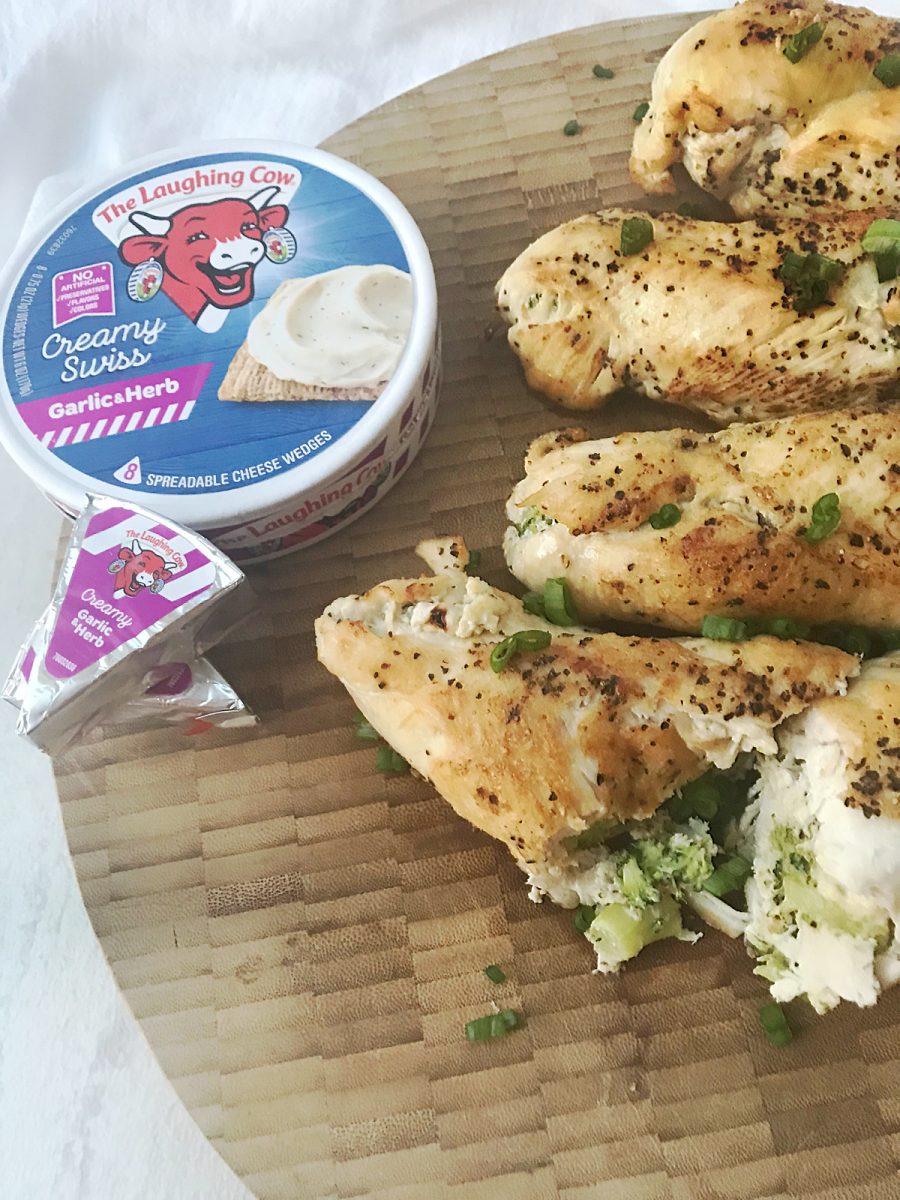 Sliced stuffed chicken rolls resting on a wooden board alongside a round container of Garlic and Herb Laughing Cow cheese.