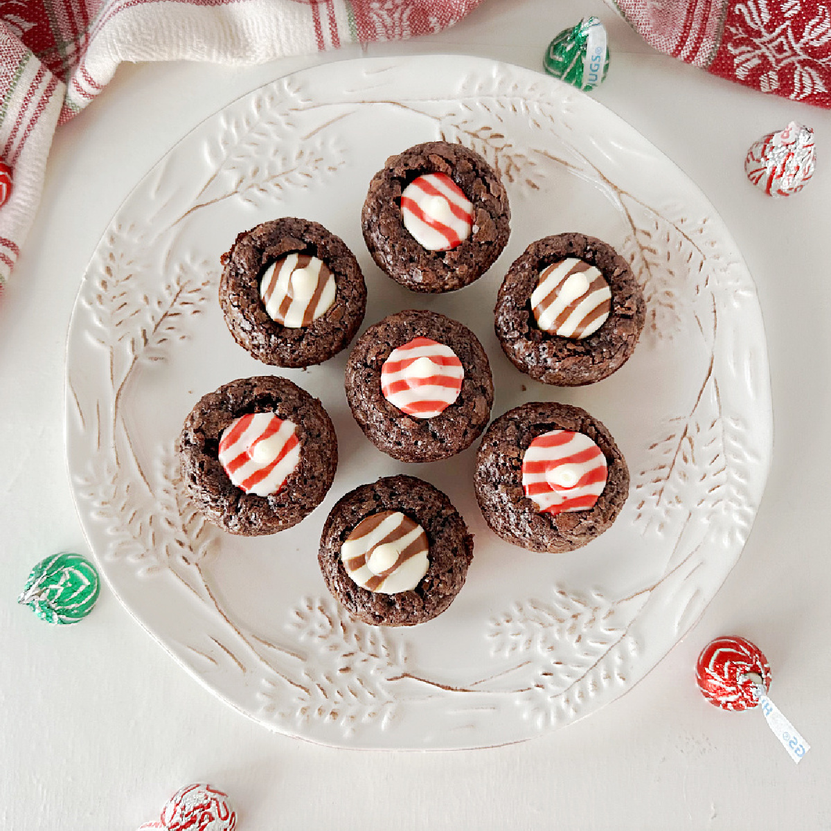 A plate of mini brownie bites, each topped with a Hershey's Kiss candy. The brownies are rich-looking, and the candies add a festive touch. The image is taken from above, and the plate is placed on a white surface.