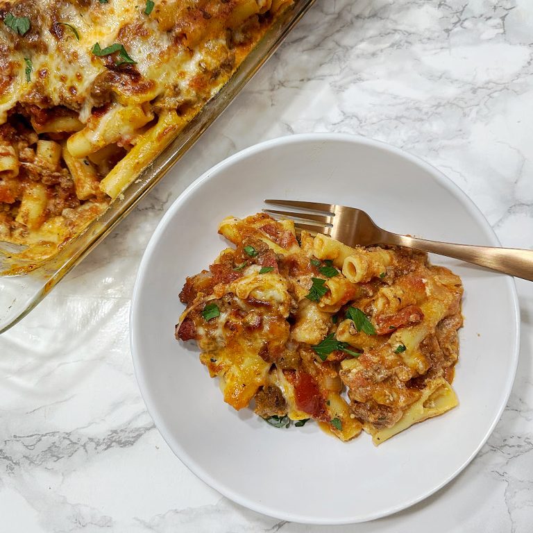 A top-down view of a delicious Baked Ziti with Ground Beef. A white bowl filled with the cheesy pasta dish sits on a marble counter, next to a baking dish with more ziti. A gold fork rests on the bowl, ready to dig in.