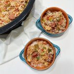 Top-down view of two blue-handled bowls filled with Crock Pot jambalaya containing rice, sausage, chicken, bell peppers, and tomatoes, next to a slow cooker with more jambalaya and a white cloth napkin underneath