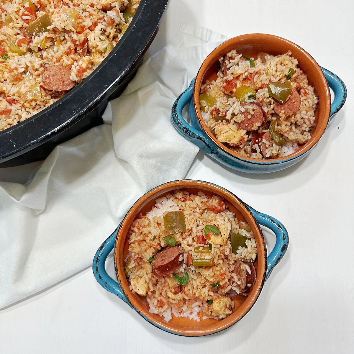 Top-down view of two blue-handled bowls filled with Crock Pot jambalaya containing rice, sausage, chicken, bell peppers, and tomatoes, next to a slow cooker with more jambalaya and a white cloth napkin underneath