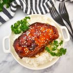 Overhead view of a serving of crock pot sticky chicken on white rice in a white handled bowl. The chicken is coated in a dark, glossy sauce and topped with sesame seeds and fresh parsley. Two black forks and a black and white striped napkin are visible on the white marble background.