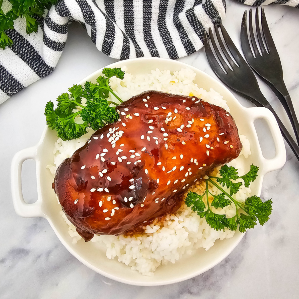 Overhead view of a serving of crock pot sticky chicken on white rice in a white handled bowl. The chicken is coated in a dark, glossy sauce and topped with sesame seeds and fresh parsley. Two black forks and a black and white striped napkin are visible on the white marble background.