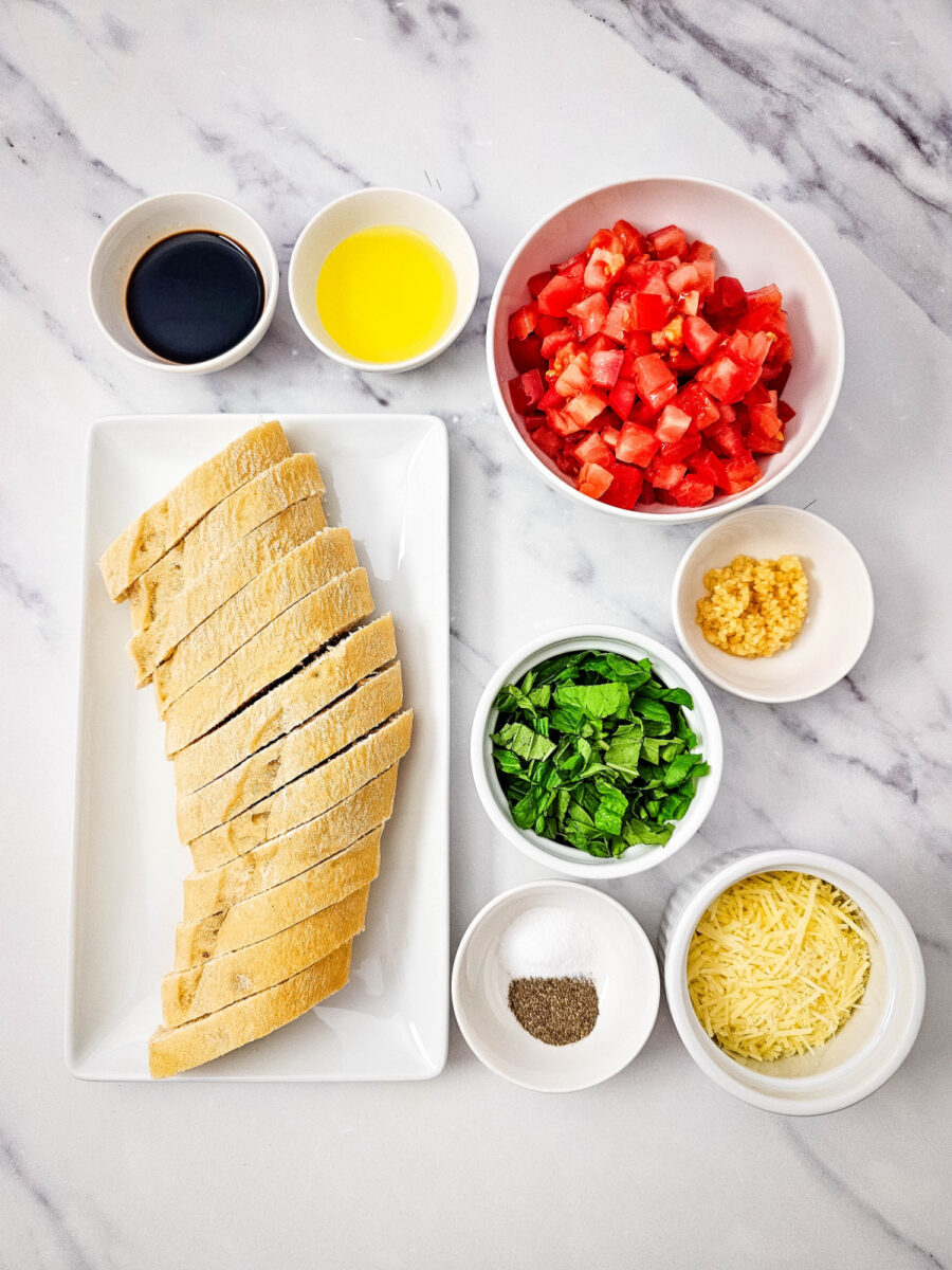 Ingredients for Bruschetta including sliced French baguette, Roma tomatoes, fresh basil, Parmesan cheese, olive oil, balsamic vinegar, garlic, salt, and pepper.