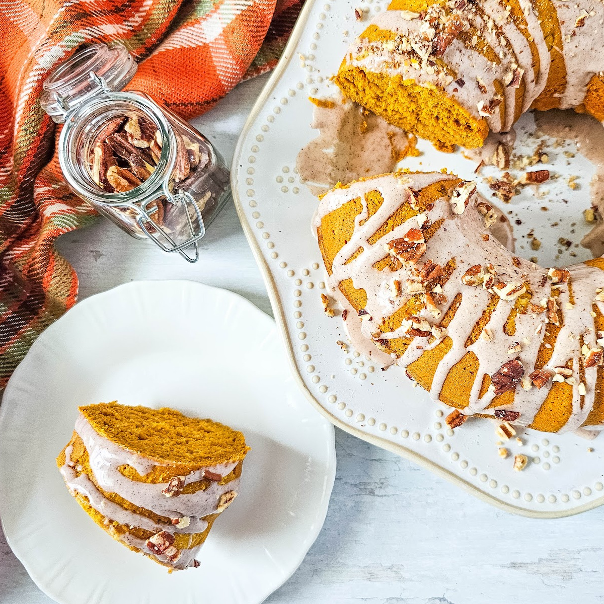 A top-down view of a pumpkin spice bundt cake on a white plate. The cake is glazed and sprinkled with pecans. A slice of the cake is on a smaller plate beside it. A jar of pecans and a folded orange and black plaid napkin are in the background.