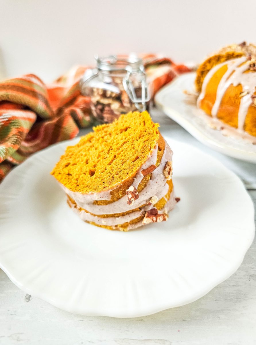 A thick slice of pumpkin bread on a white plate, drizzled with white glaze and topped with a pecan. The bread is a vibrant orange color and appears moist.