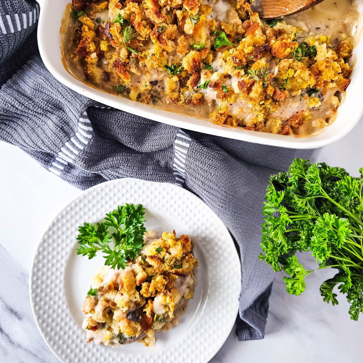 Overhead view of a rectangular baking dish filled with golden brown stuffing, with a wooden spoon inside. A white plate with a portion of stuffing garnished with parsley sits next to the dish, along with a bundle of fresh parsley.