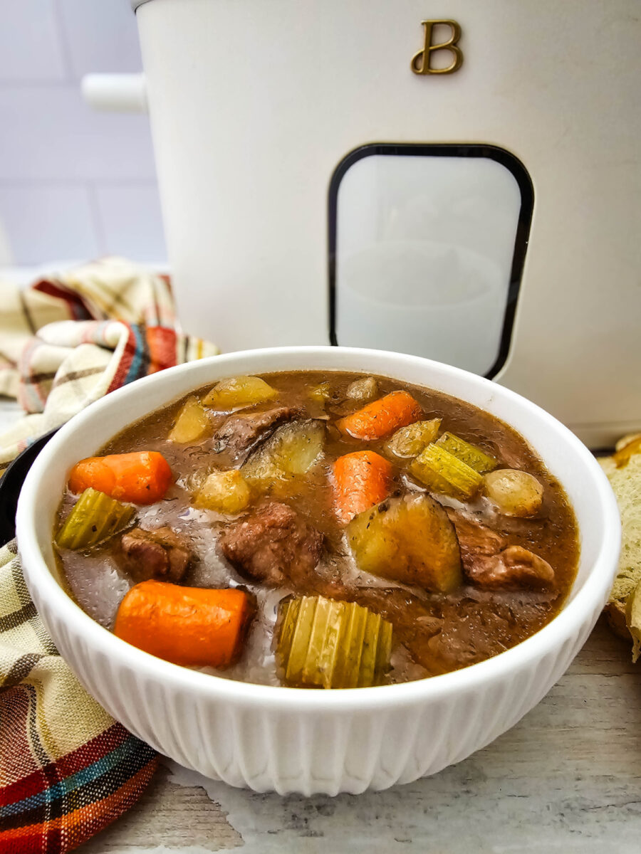 Easy Crock Pot Beef Stew served in a white bowl with chunks of beef, potatoes, carrots, celery, and pearl onions in rich brown gravy, with a slow cooker in the background.