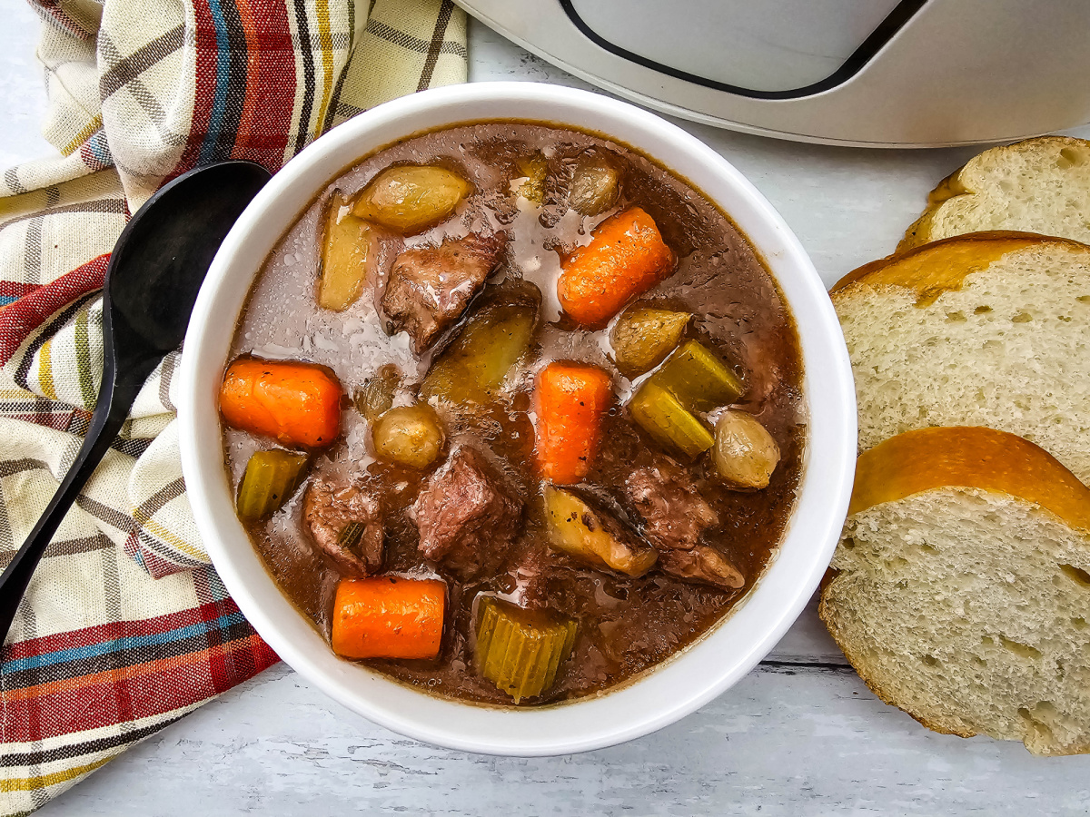Overhead view of a bowl of beef stew with tender beef, potatoes, carrots, celery, and pearl onions, served with sliced bread and a plaid kitchen towel.