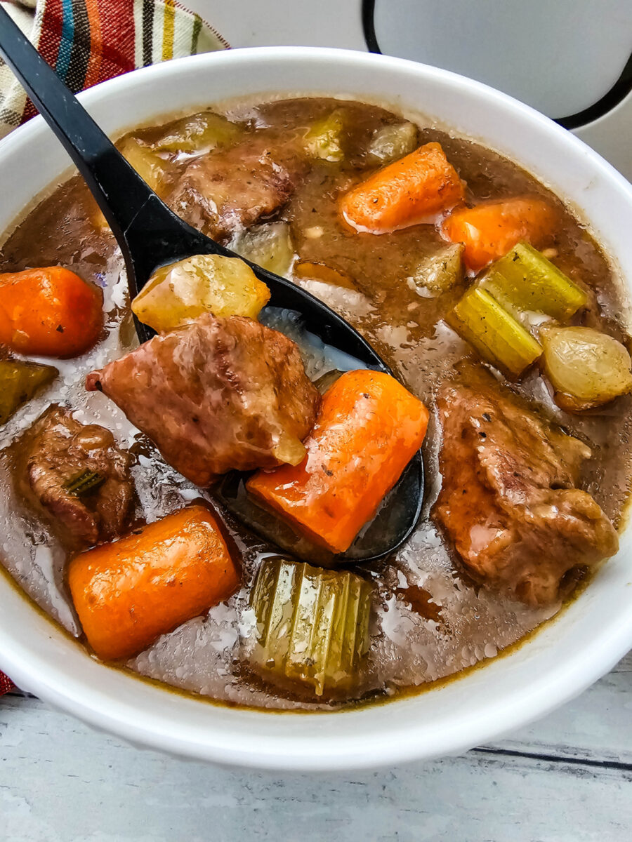 Close-up of beef stew in a white bowl with a black spoon holding a piece of beef, potato, and carrot above the rich gravy and vegetables.