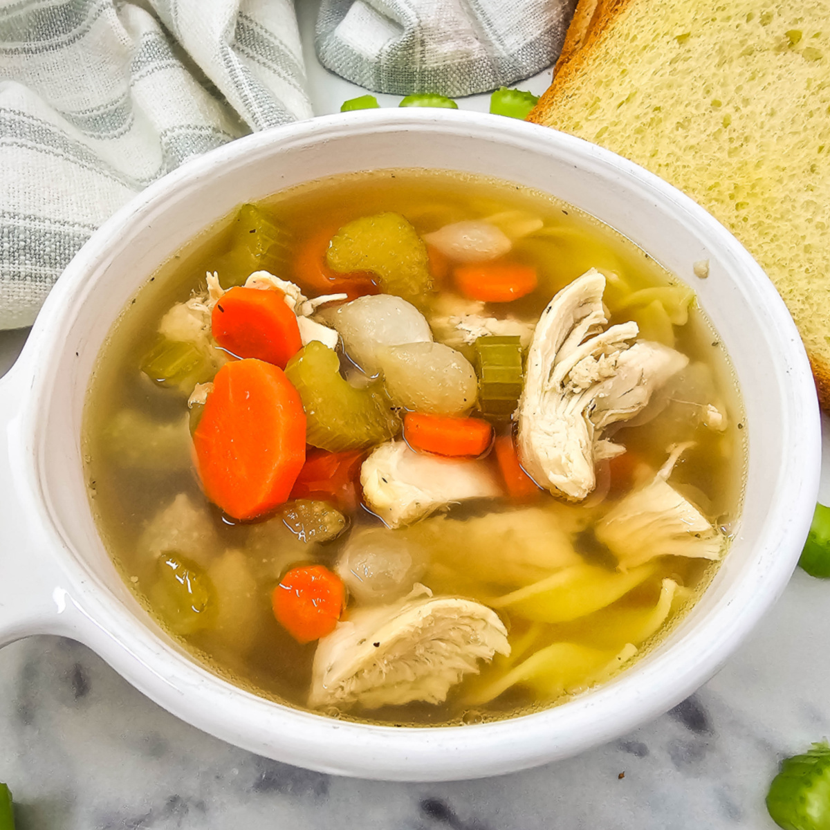 Bowl of chicken noodle soup with chicken, carrots, celery, and noodles. A slice of bread is on the side. The bowl sits on a marble surface with a patterned cloth in the background.