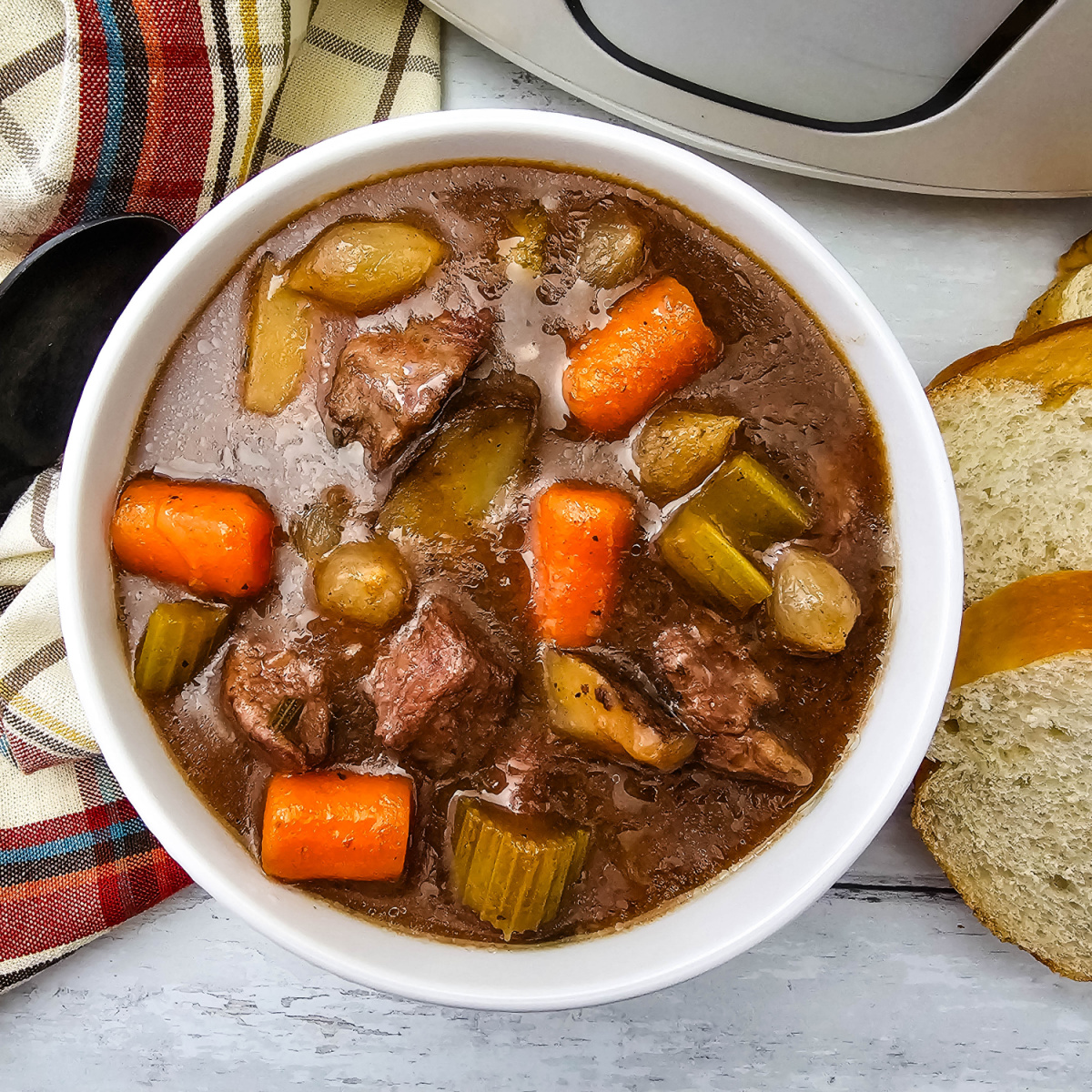 Overhead view of Easy Crock Pot Beef Stew in a white bowl with beef, potatoes, carrots, celery, and pearl onions, with sliced bread and a plaid towel beside it.