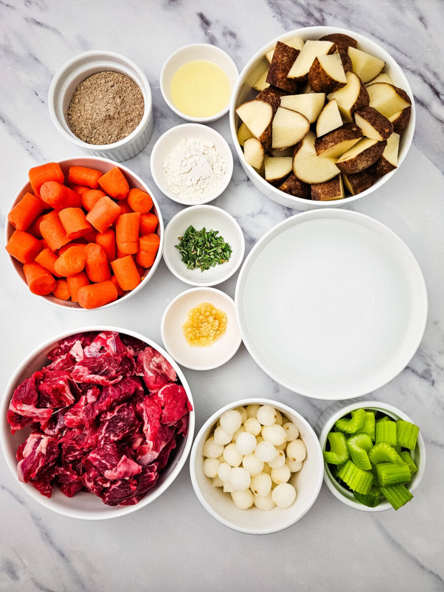 Overhead view of the ingredients for Crock Pot Beef Stew arranged in bowls, including beef, potatoes, carrots, celery, pearl onions, rosemary, garlic, flour, oil, seasoning mix, and water.