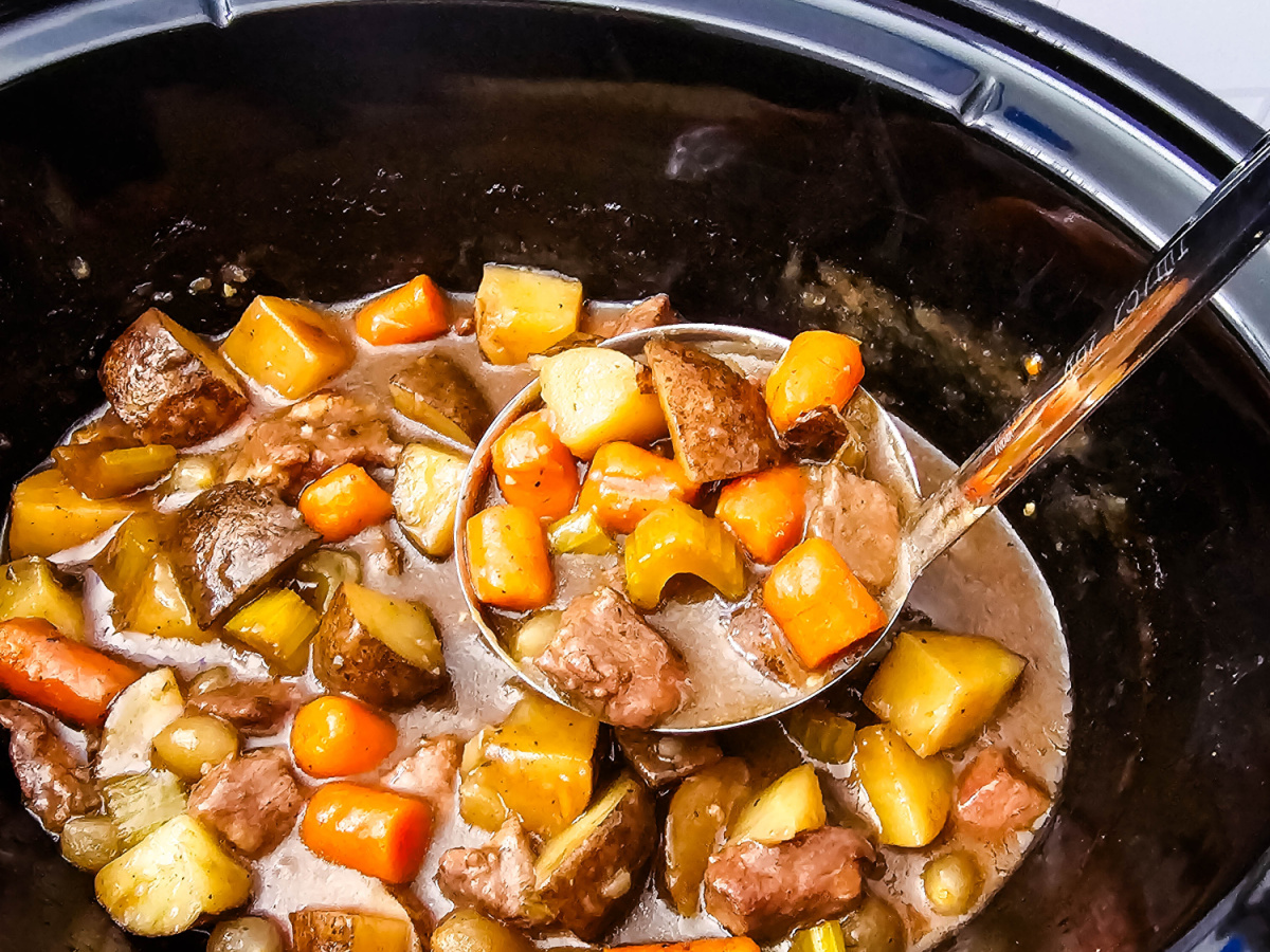 Close overhead view of cooked beef stew inside a slow cooker with a ladle holding beef, potatoes, carrots, and celery in the gravy.