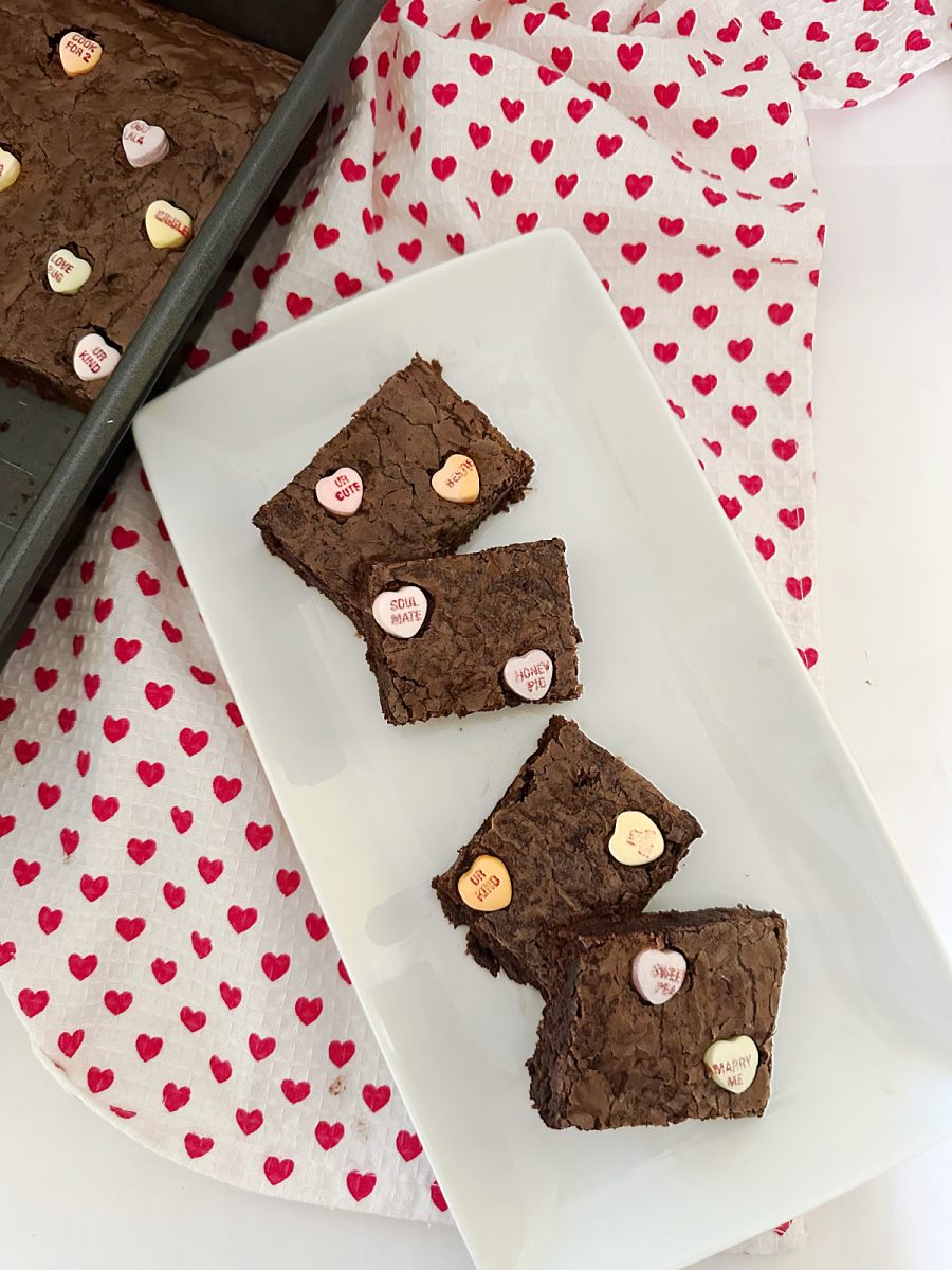 A high-angle shot showing four fudgy treats topped with colorful conversation hearts on a white platter. To the left, a portion of the baking pan and a red heart-patterned kitchen towel are visible.