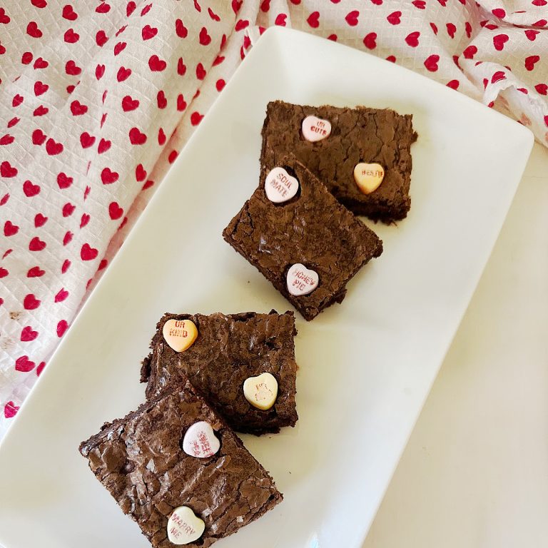 An overhead shot of four thick, chewy cocoa bars topped with candies, positioned diagonally on a white plate. A heart-patterned cloth is draped in the background to highlight these seasonal sweets.