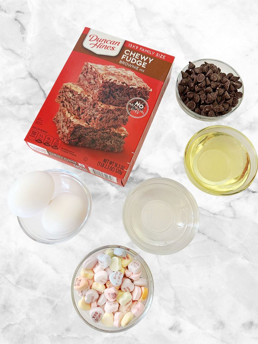 An overhead shot of components for this recipe on a marble surface. The items include a box of Duncan Hines mix, two eggs, chocolate chips, vegetable oil, water, and a bowl of colorful heart-shaped candies.