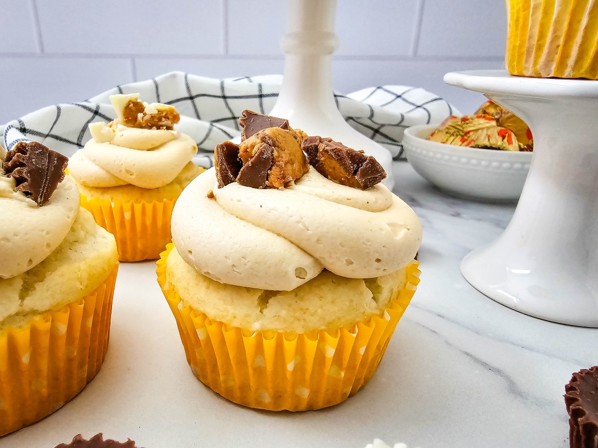 Close-up of a cupcake with a thick swirl of peanut butter buttercream and chopped chocolate peanut butter cups, with more cupcakes behind it.