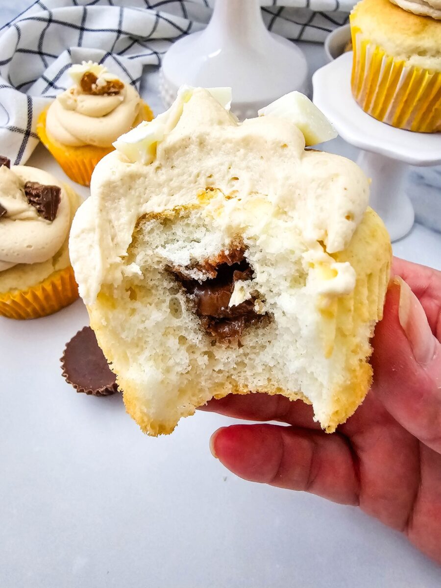 A hand holds an opened vanilla cupcake, revealing a chocolate peanut butter cup baked into the center beneath a thick layer of frosting.