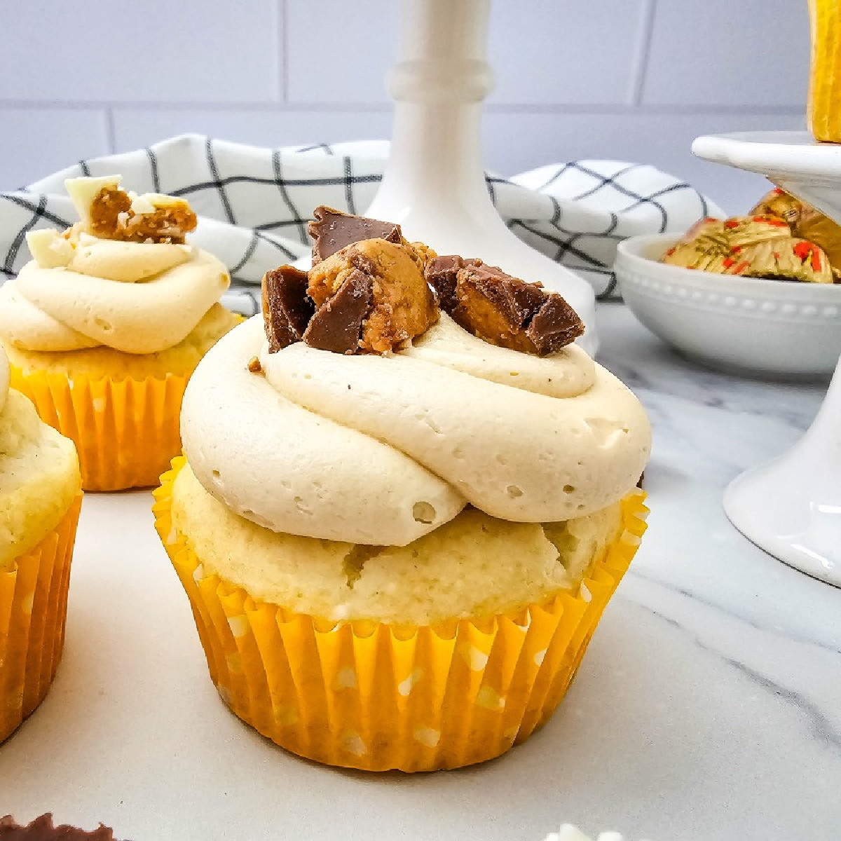 Vanilla Peanut Butter Cup Cupcakes in yellow liners, with a close-up of piped peanut butter frosting and chopped chocolate candy on the front cupcake.