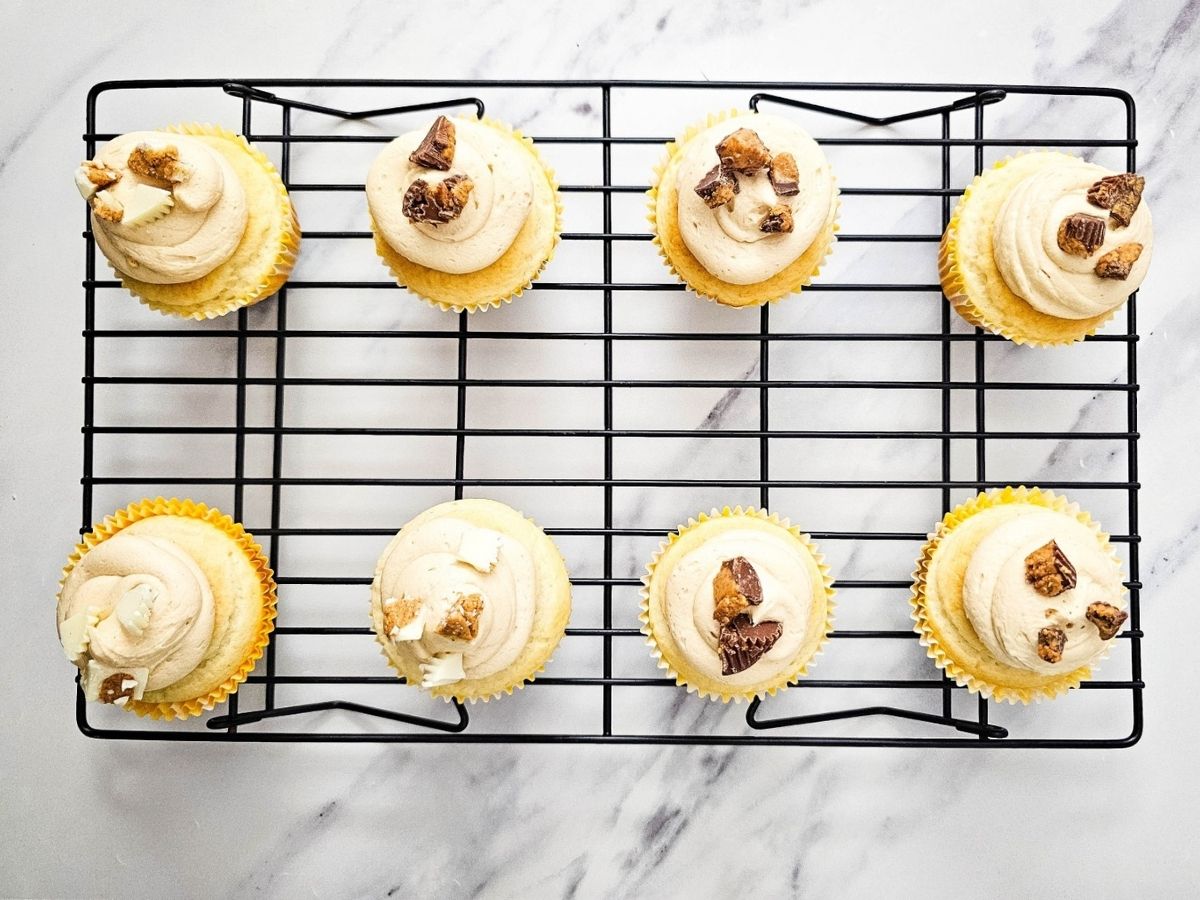 Eight cupcakes on a black wire rack, each decorated with a swirl of peanut butter frosting and pieces of milk chocolate or white chocolate candy.