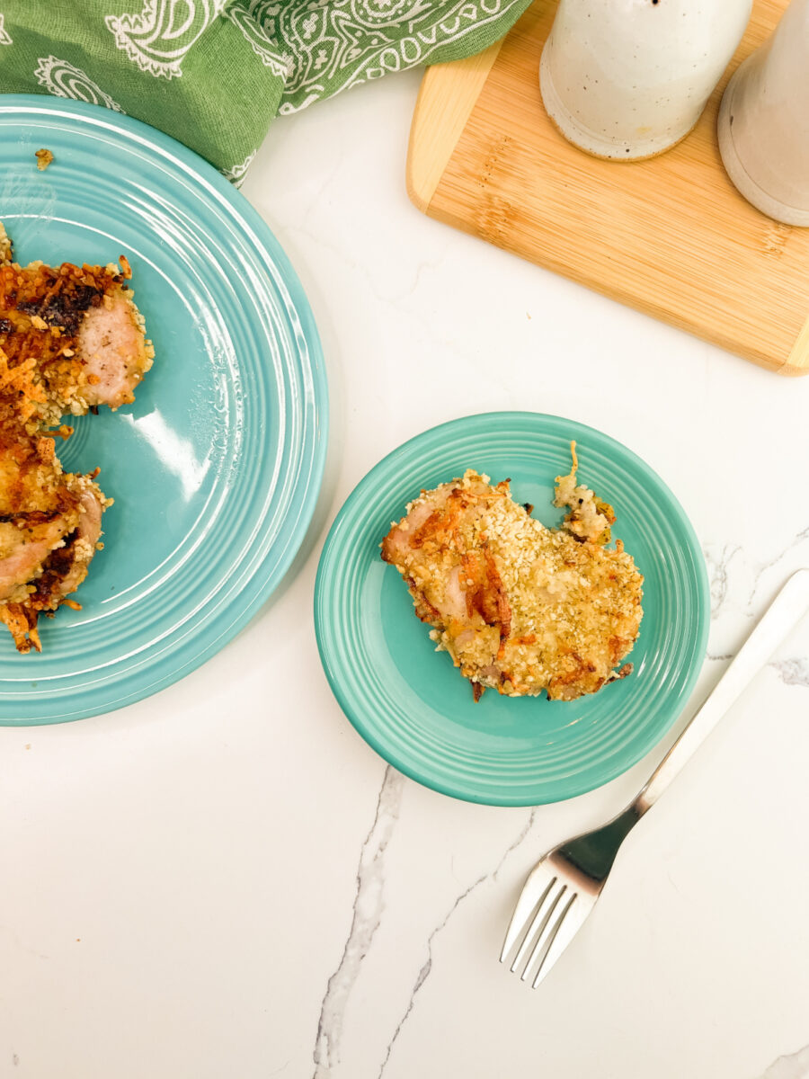 Breaded pork chop on a turquoise plate with a platter of additional pork chops in the background on a white marble countertop.