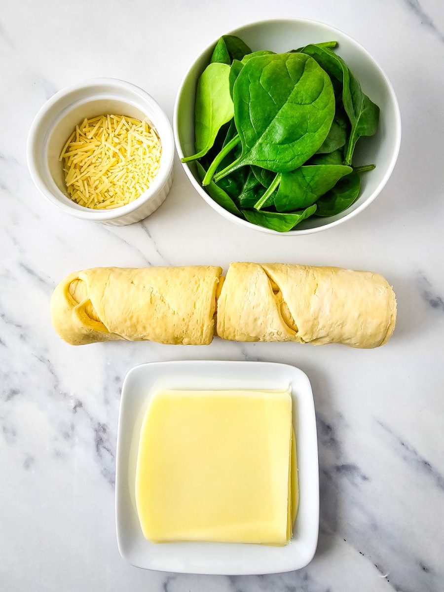 An overhead flat lay of the ingredients for the recipe arranged on a marble surface, including a tube of crescent roll dough, a bowl of baby spinach, a bowl of shredded Parmesan cheese, and a square of slices mozzarella cheese.