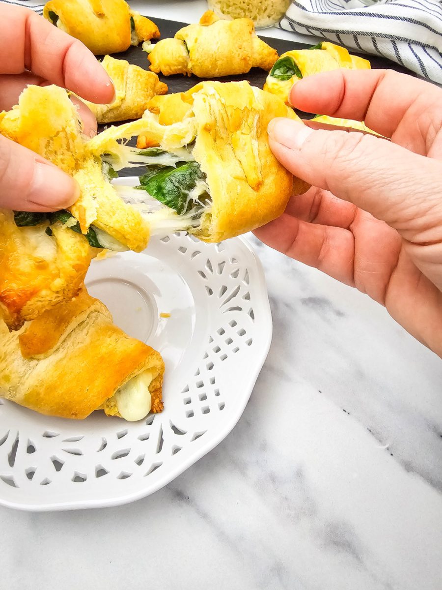 A close-up shot of two hands pulling a baked spinach and mozzarella crescent roll apart, revealing the gooey, stretchy melted cheese and green spinach inside.