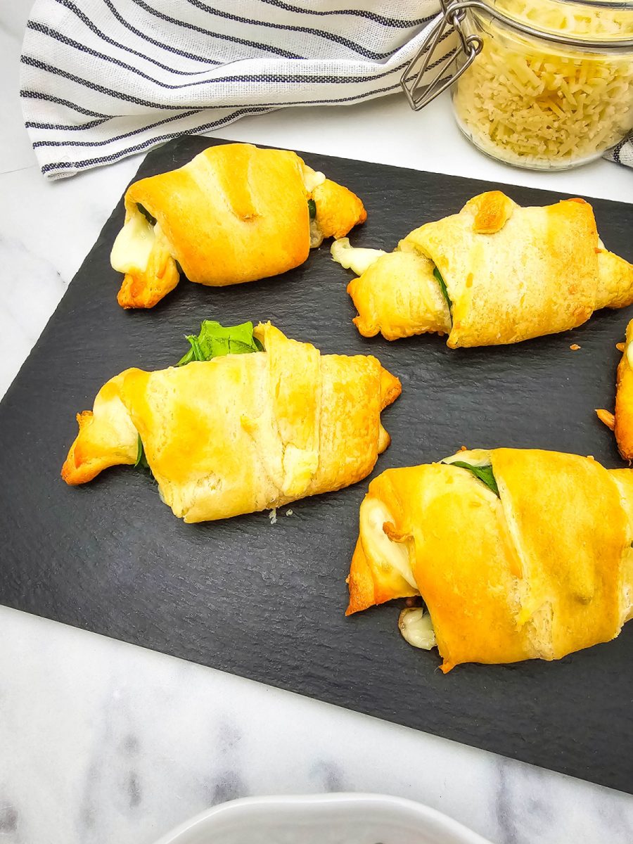 A collection of baked spinach and cheese crescent rolls are arranged on a black slate board, with a glass jar of shredded cheese and a striped napkin in the background.