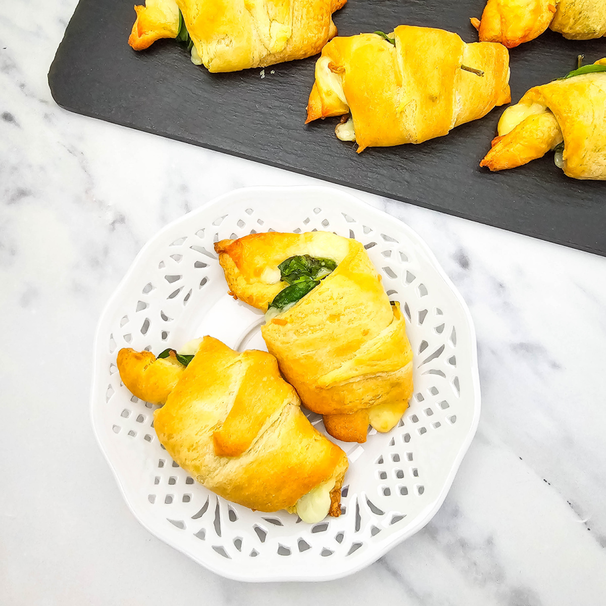 Two golden-brown spinach and cheese crescent rolls are featured on a decorative white plate, with a black slate board of more rolls in the blurred background.