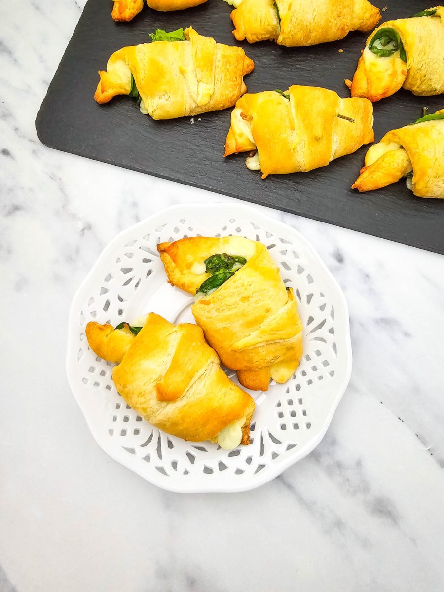 An overhead shot of baked spinach and cheese crescent rolls. Two rolls are on a white decorative plate in the foreground, with more rolls on a black slate board in the background.