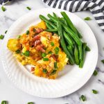 Overhead view of a white plate with a serving of tater tot casserole with visible chunks of tater tots, cheese, and bacon bits. Green beans are arranged neatly beside the casserole. The plate is on a marble surface with scattered green onion pieces and a striped napkin.