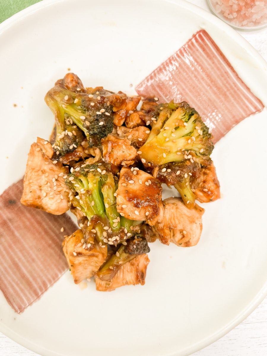 Chicken and broccoli stir-fry coated in a savory sauce and sprinkled with sesame seeds, served on a white plate with a decorative brown and white stripe.