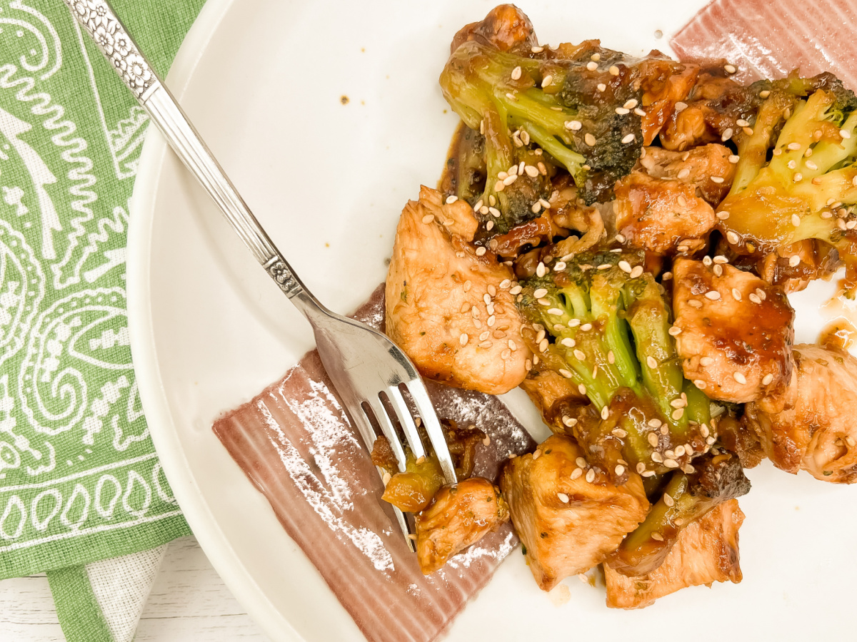 A fork rests on a plate of easy chicken and broccoli skillet meal, showcasing tender chicken pieces and vibrant green broccoli florets coated in sauce, with a green patterned napkin to the side.