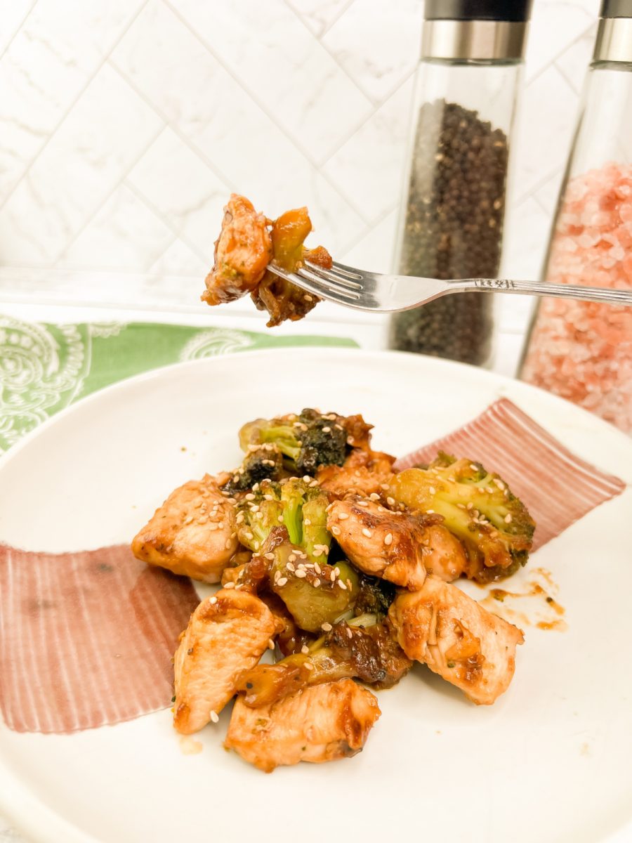 A fork holds a bite of chicken and broccoli over a plate filled with the chicken and broccoli stir-fry, with salt and pepper grinders blurred in the background.