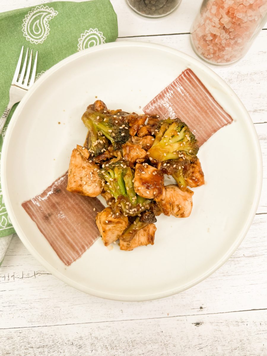 An overhead view of a one-pan chicken and broccoli recipe on a white plate, garnished with sesame seeds, alongside a green napkin and a fork.