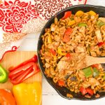 An overhead shot of a large skillet filled with one pan chicken fajita pasta next to a wooden cutting board with sliced fresh bell peppers.