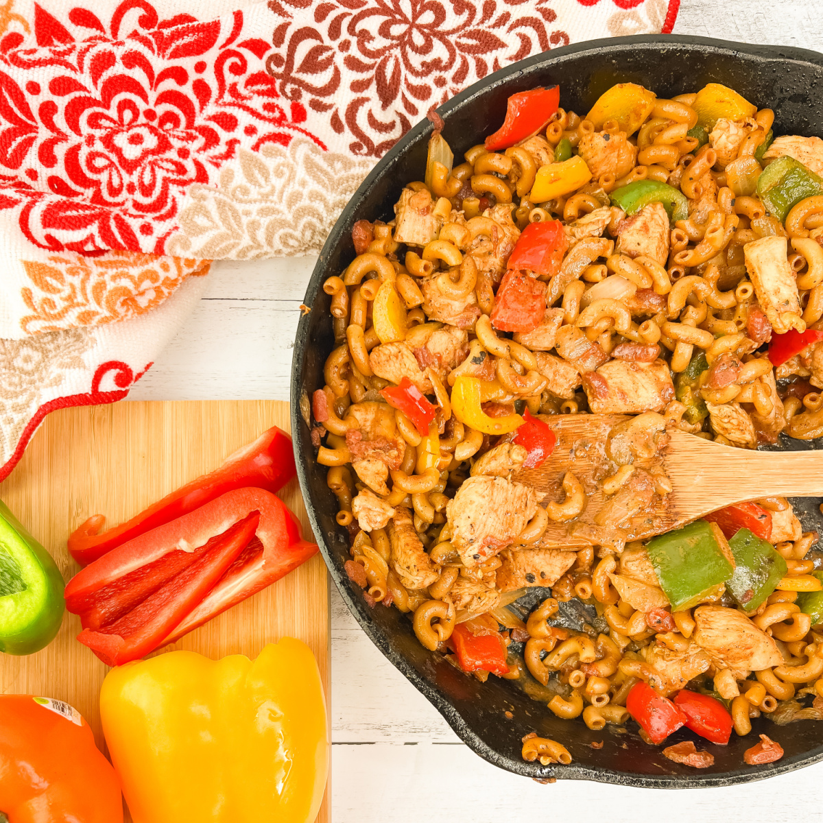 An overhead shot of a large skillet filled with one pan chicken fajita pasta next to a wooden cutting board with sliced fresh bell peppers.