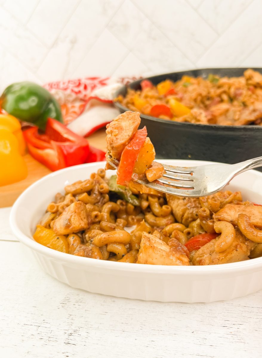 fork holds a perfect bite of seasoned chicken, a red bell pepper slice, and elbow macaroni over a bowl of creamy pasta.