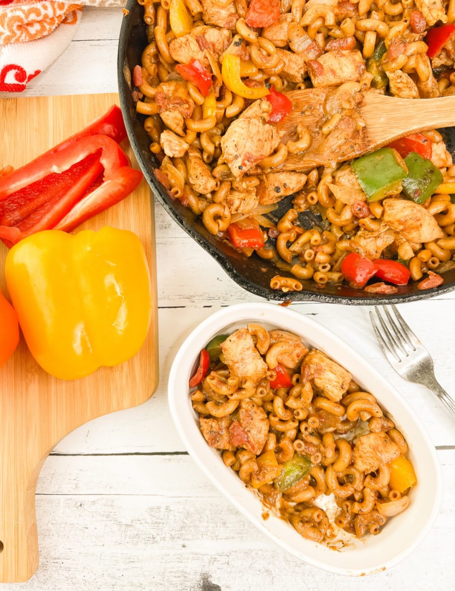 A wide overhead view showing a cast iron skillet and a small white serving bowl both filled with a zesty chicken and vegetable pasta dish.