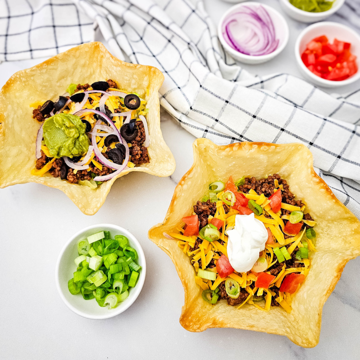 Easy Taco Salad Bowls with ground beef, lettuce and cheddar in golden tortilla shells, one topped with guacamole and the other with sour cream.