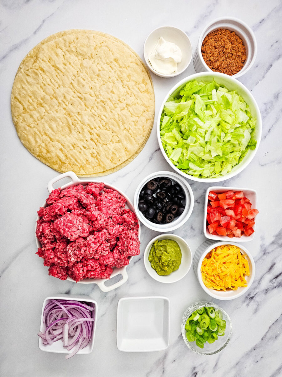 flour tortillas, ground beef, taco seasoning, lettuce, cheddar, tomatoes, onions, olives, sour cream and guacamole arranged in bowls.